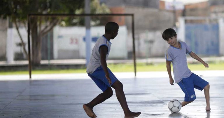 Descrição de Imagem: Fotografia de dois meninos em uma quadra jogando futebol, eles vestem um uniforme escolar com camiseta branca e bermuda azul. Um dos meninos possui deficiência física de membro inferior e não possui os pés.