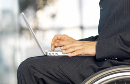 Descrição de Imagem: Foto de um homem em cadeira de rodas vestindo terno e digitando em um notebook que está em seu colo.
