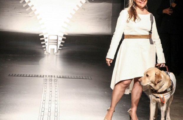 Descrição de Imagem: A fotografia mostra uma modelo cega com um cão guia, desfilando na passarela. Ela veste um vestido branco de cumprimento acima dos joelhos na frente e abaixo dos joelos atrás. Um cinto marron na cintura e mangas compridas. Na passarela um grande zíper aberto serve de cenário.