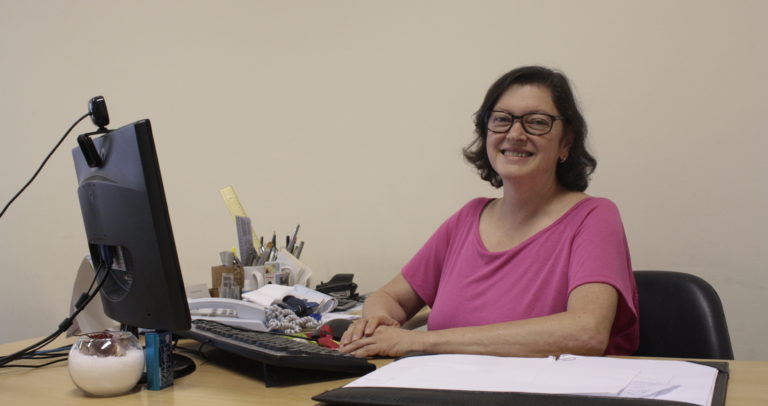 Descrição da Imagem: Fotografia de Valquiria, uma mulher que usa óculos e tem cabelos escuros na altura do ombro. Está sentada em sua mesa de trabalho que possui um computador e papéis. Veste uma camiseta cor de rosa e está sorrindo.