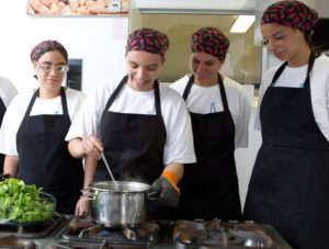 Descrição da Imagem: Fotografia de uma turma de aprendizes da APAE. Quatro mulheres estão em uma cozinha vestindo avental preto e toucas. Uma delas mexe em uma panela que está no fogão com colher de pau enquanto as outras observam. Todas estão sorrindo.