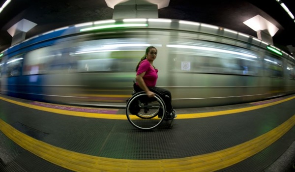Descrição de Imagem: Mulher cadeirante em uma plataforma do metrô com o trem passando ao fundo.