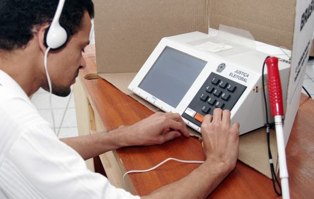 Descrição de Imagem: Um eleitor cedo sentado em uma cabine de votação. Está usando fones de ouvido ao utilizar a urna eletrônica.