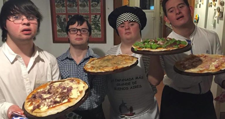 Descrição de Imagem: Fotografia dos quatro pizzaiolos com Síndrome de Down. São todos homens jovens vestidos com camisa social e avental. No centro um deles está usando um chapéu de cheff de cozinha. Cada um segura uma pizza de sabores diferentes.