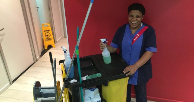 Descrição de Imagem: Fotografia de Georgina no ambiente de trabalho. Georgina é uma mulher negra e está sorrindo. Veste um uniforme azul com logo da Sodexo na camisa e está ao lado de um carrinho de limpeza amarelo que contém utensílios e produtos de limpeza