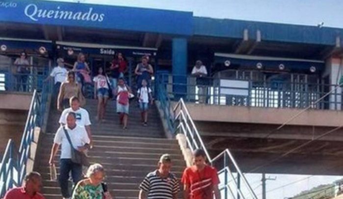Descrição de Imagem: Fotografia da entrada da estação d etrem de Queimados no Rio de Janeiro. Mostra uma grande escada e pessoas entrando e saindo da estação.