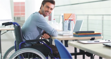 Descrição de Imagem: Homem cadeirante com os braços apoiados em sua mesa de trabalho. É um homem branco com cabelos escuros curtos e barba por fazer. Está sorrindo e vestes uma blusa azul clara de mangas compridas. Sobre a mesa estão alguns livros e um laptop.