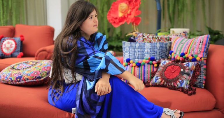Descrição de Imagem: Fotografia de Isabella Springmuhl, estilista com Sindrome de Down. Ela está sentada com as pernas cruzadas em um sofá laranja rodeada de várias almofadas coloridas. Tem os cabelos castanhos e bem compridos, veste um vestido longo em vários tons de azul.
