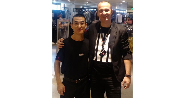 Descrição de Imagem: Fotografia de Dan Fukushima, trabalhador com deficiência física, nas lojas Renner. Dan é oriental, tem cabelos bem curtos e escuros e usa óculos.Veste um uniforme com camisa e calça pretas. Ao seu lado direito está o coordenador da loja em que trabalha, um homem branco de cabelos curtos mais alto que Dan. Os dois estão sorrindo.