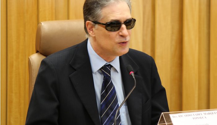 Descrição de Imagem: Fotografia do desembargador Ricardo Tadeu. Está sentado em uma cadeira de couro marrom atrás de uma mesa. É um homem com cabelos grisalhos e usa óculos escuros, veste um paletó e gravata escuros e camisa azul clara.
