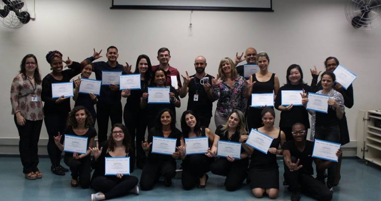 Descrição de Imagem: Fotografia dos 17 participantes da décima quarta Oficina de Surdez e Acessibilidade promovida pelo CEPRO. Todos os formados pela Oficina estão vestidos de preto e segurando o certificado nas mãos. Com a outra mão fazm o sinal de I Love You (Eu amo você em inglês) na língua de sinais. No centro da foto está Susana Penteado, coordenadora do CEPRO, e à esquerda está Fernanda Oliveira, técnica do CEPRO.