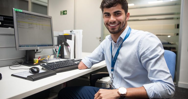 Descrição de Imagem: Fotografia de Ricardo Vieira. Está sentado em sua mesa de trabalho, é um homem jovem com cabelos castanhos e barba curta, tem o antebraço direito amputado. Veste uma camisa social azul e calça escura, está sorrindo.