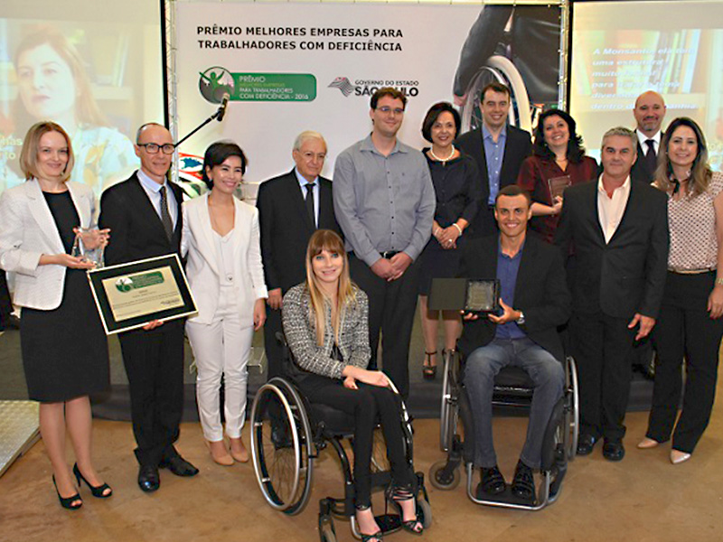 Descrição de Imagem: Fotografia tirada no evento de premiação. Na fotografia estão os organizadores do evento e reprentantes das empresas premiadas. Na frente estão dois cadeirantes, um homem e uma mulher. Na fileira de trás estão sete pessoas, três mulheres e quatro homens. Todos estão vestidos com roupa social e sorrindo. Alguns seguram as placas de premiação distribuídas para as empresas vencedoras. Ao fundo está um painel branco onde está escrito PRÉMIO MELHORES EMPRESAS PARA PESSOAS COM DEFICIÊNCIA, Governo do Estado de São Paulo