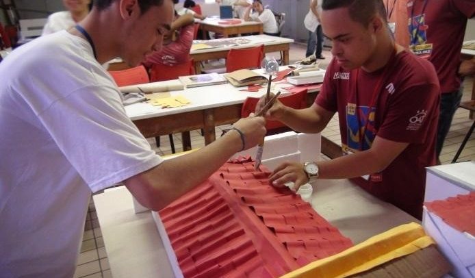 Descrição de Imagem: Fotografia da oficina da OAT com participantes do Programa Expressão Artística. Mostra um profissional da OAT pintando o telhado da uma maquete de uma casa junto com um jovem atendido pela instituição. Os dois seguram pinceis e pintam o telhado de laranja.