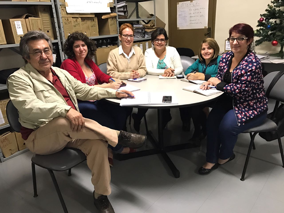 Descrição de Imagem: Fotografia tirada na reunião da Secretaria Administrativa realizada em uma sala na SRTE-SP. Os membros estão sentados ao redor de uma mesa redonda, na mesa estão alguns papéis e canetas. No canto direito está uma árvore de natal e ao fundo duas estantes com algumas caixas de papelão que guardam arquivos.. Todos estão sorrindo.