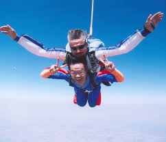 Descrição de Imagem: Fotografia de Ricardo pulando de paraquedas acompanhado de um instrutor. Mostra os dois em queda livre com os braços abertos, ambos estão sorrindo.