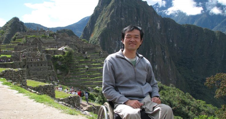 Descrição de Imagem: Fotografia de Ricardo em Machu Pichu. Ele é um homem oriental com cabelos curtos, pretos e lisos. Está de cadeira de rodas e veste um moletom cinza. Ao fundo estão montanhas, escadarias e gramados de Machu Pichu, está um dia ensolarado.