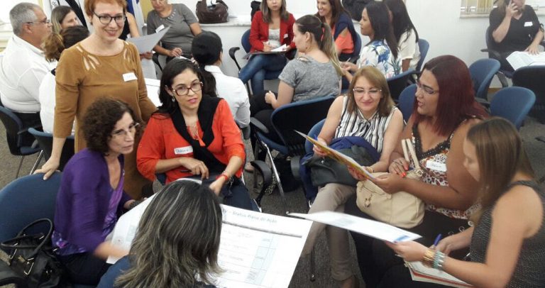 Descrição de Imagem: Fotografia de gestores reunidos em círculos debatem materiais que têm em mãos sobre gestão para a inclusão. Numa das rodas Ivone Santana, do Instituto Modo Parités, está em pé, de blusa cor de mostarda, orientando as tarefas.