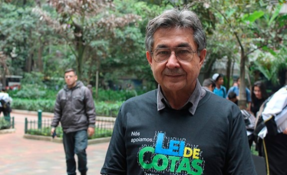 Descrição de Imagem: Fotografia do Dr.Kal, um homem com cabelos curtos e grisalhos que usa óculos. Ele veste uma camiseta preta comemorativa do aniversário da Lei de Cotas, está em uma praça e atrás dele estão muitas árvores.