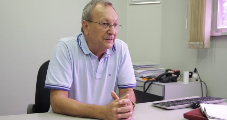 Descrilção de Imagem: Fotografia de Osmar Spinussi sentado em sua mesa de trabalho. Osmar é um homem branco, com cabelos grisalhos e óculos. Está com as mãos apoiadas sobre a mesa e veste uma camisa polo branca.