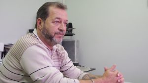 Descrição de Imagem: Fotografia de Romualdo Siviera, um homem branco com cabelo e barbas grisalhos. Está sentado em uma mesa de trabalho e veste uma blusa de lã rosa com listras em marrom