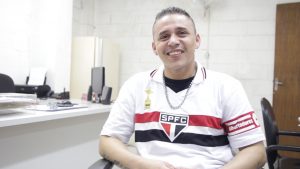 Descrição de Imagem: Fotografia de Francisco, homem branco, jovem, com cabelo curto. Está sentado em uma cadeira e veste uma camiseta do São Paulo Futebol Clube. Está sorrindo.