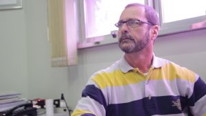 Descrição de Imagem: Fotografia de Adalberto, homem branco com cabelos e barba grisalhas, veste uma camisa polo com litras amarelas e azuis e usa óculos.