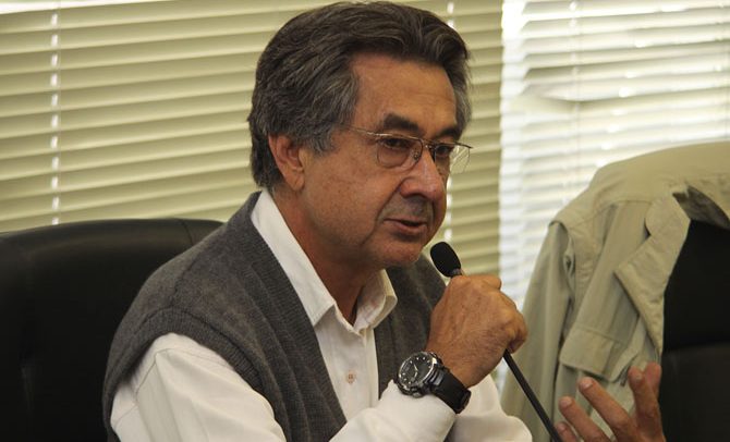 Descrição de Imagem: Fotografia do Dr. José Carlos do Carmo, conhecido como Kal. É um homem com cabelos curtos e grisalhos e usa óculos Ele está palestrando e segurando um microfone, vesta uma camisa branca de manga comprida e um colete de lá cinza.
