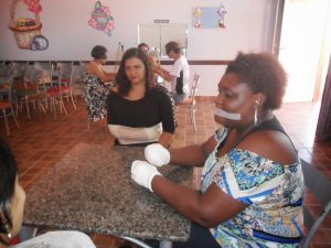 Descrição de Imagem: Fotografia de uma oficina realizada pela APARU. Mostra uma sala com pessoas conversando reunidas em duas mesas, na mesa mais a frente estão três mulheres, duas delas estão com braços e mãos enfaixados e amarrados, simulando uma limitação física.