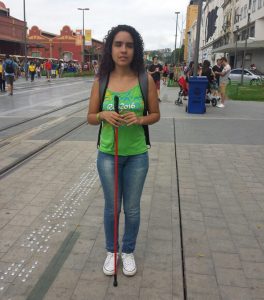 Descrição de Imagem: Fotografia de Vitória Maria, mulher de cabelos cacheados e cega. Está de pé em uma calçada com piso tátil segurando sua bengala vermelha com as duas mãos, veste uma regata verde e calça jeans.