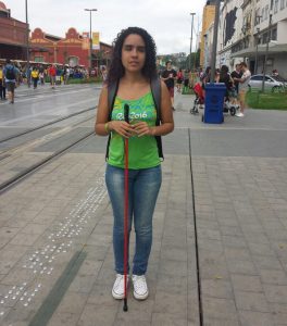 Descrição de Imagem: Fotografia de Vitória Maria, mulher de cabelos cacheados e cega. Está de pé em uma calçada com piso tátil segurando sua bengala vermelha com as duas mãos, veste uma regata verde e calça jeans. 