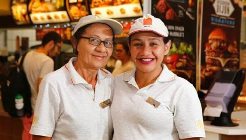 Descrição de Imagem: Fotografia de mãe e filha funcionárias do McDonalds. Elas estão abraçadas e vestem o uniforme do McDonals com camisetas e bonés brancos com o logo da lanchonete, um M amarelo. Ambas estão sorrindo.