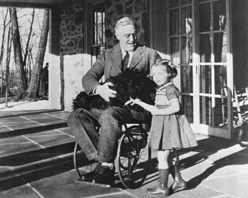 Descrição de Imagem: Fotografia em preto e branco do ex-presidente Franklin Roosevelt. Na fotografia ele já está idoso, tem cabelos brancos e está em uma cadeira de rosas, veste um terno escuro e usa óculos. Está na frente de uma casa, em seu colo está um cachorro preto e ao seu lado está uma menina de cabelos curtos e vestido com aparência de 6 anos. Todos estão sorrindo