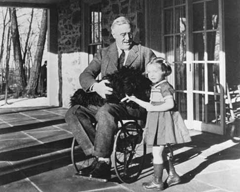 DescriÃ§Ã£o de Imagem: Fotografia em preto e branco do ex-presidente Franklin Roosevelt. Na fotografia ele jÃ¡ estÃ¡ idoso, tem cabelos brancos e estÃ¡ em uma cadeira de rosas, veste um terno escuro e usa Ã³culos. EstÃ¡ na frente de uma casa, em seu colo estÃ¡ um cachorro preto e ao seu lado estÃ¡ uma menina de cabelos curtos e vestido com aparÃªncia de 6 anos. Todos estÃ£o sorrindo