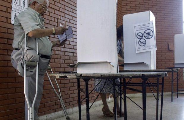 Descrição de Imagem: Fotografia de um homem com deficiência física diante de uma cabine eleitoral. É o homem idoso, negro e de cabelos brancos, usa duas muletas e possui a perna direita amputada.
