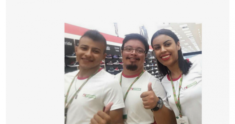 Arthur está ao lado de dois colegas de trabalho, um rapaz e uma moça. Todos estão de camiseta e crachá da loja de esportes Centauro, onde trabalham, fazendo sinal positivo. Arthur é o único da foto com síndrome de Down.