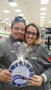 De uniforme cinza da marca esportiva Fila e com o logotipo da Centauro, Arthur recebe uma caixa de bombons da gerente da loja. Ela também usa o mesmo uniforme que ele. Cada um segura os bombons com uma mão. Arthur tem um óculos de sol na testa e usa bigodes e cavanhaque. Ela é loira e usa óculos. Eles sorriem. e ao fundo se vê o interior da loja.