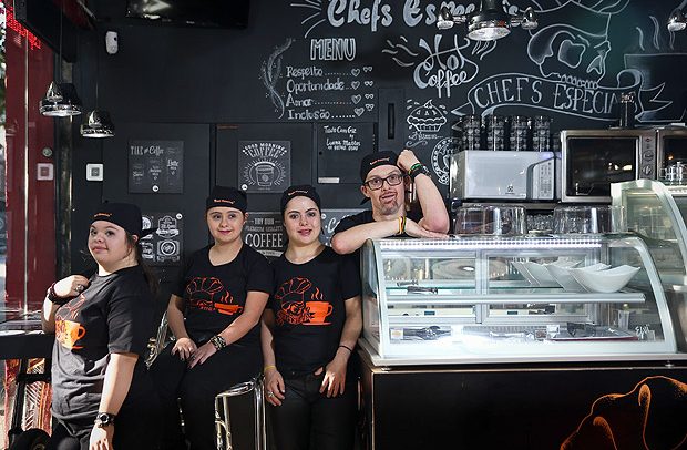 Descrição da imagem: Na frente do balcão da cafeteria, quatro atendentes: Luiza (esq.), Dani, Carol e Rodrigo, que está apoiado na vitrine de salgados. Os atendentes do novo café na região dos Jardins usam bandana preta na cabeça e uma camiseta preta com desenho em laranja do logotipo