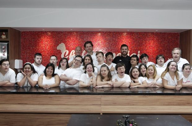 Descrição da imagem: Atrás de uma grande bancada de cozinha, estão apoiados sobre o mármore preto, dezenas de jovens que participam do programa Chefs Especiais. Todos vestem camiseta branca. Há rapazes e moças. Ao fundo alguns chefs renomados da culinária e também os idealizadores do Instituto, Simone e Márcio Berti