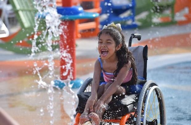 Descrição da imagem: uma menina de cerca de 10 anos na cadeira de rodas brinca numa fonte de águas em meio a brinquedos coloridos laranja e azul. Ela sorri debruçada na cadeira tocando a água com os pés e com as mãos.    