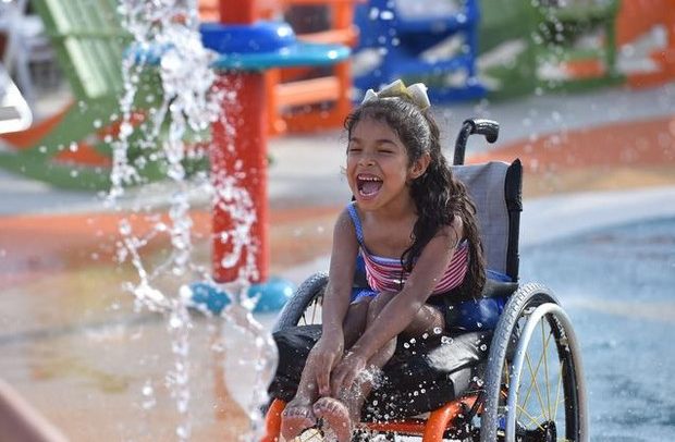 Descrição da imagem: uma menina de cerca de 10 anos na cadeira de rodas brinca numa fonte de águas em meio a brinquedos coloridos laranja e azul. Ela sorri debruçada na cadeira tocando a água com os pés e com as mãos.
