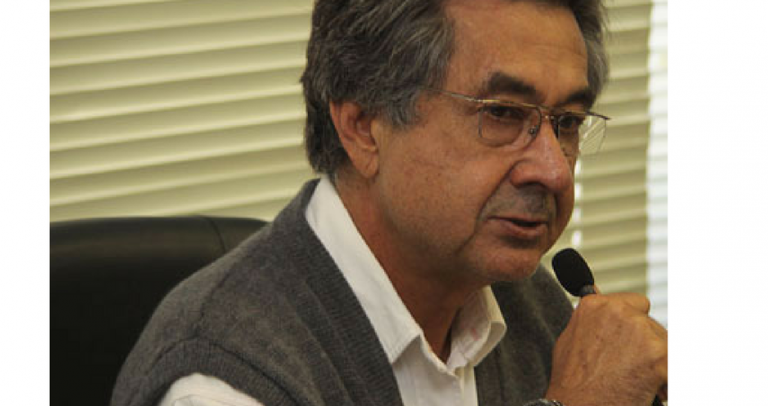 Ao microfone, dr. Kal usa óculos, tem fartos cabelos lisos e meio grisalhos. Usa colete cinza, camisa social clara e relógio de pulso preto. Ele segura o microfone com uma mão e gesticula com a outra.