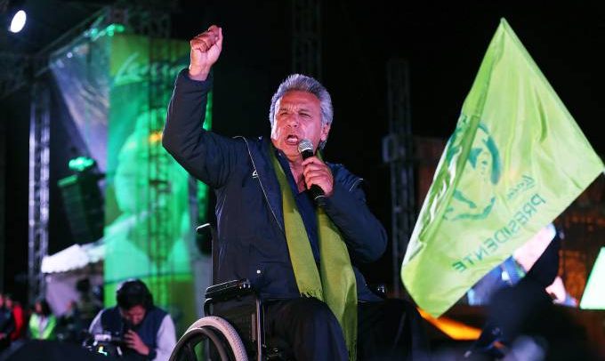 De terno escuro, echarpe verde sobre o terno e na cadeira de rodas, Lenin Moreno tem os cabelos grisalhos, a pele clara e está com a mão direita erguida e punho fechado. Bandeiras verdes tremulam em sua frente e luzes verdes iluminam o palco.