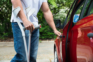 Descrição da imagem: Utilizando bengalas, motorista abre a porta de um carro vermelho. No vidro dianteiro se vê o adesivo de pessoa com deficiência
