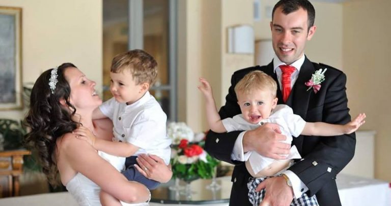 Descrição da imagem: Os pais estão vestidos de noivos - ela de branco e ele com trajes escoceses, e cada um segura um filho no colo. O que está no colo do pai parece chorar e tenta sair do colo. O que está nos braços da mãe sorri para ela.