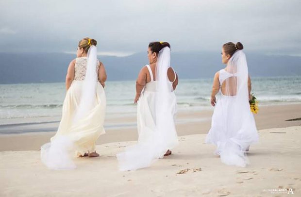 Três modelos vestidas de noivas, todas com nanismo, estão em frente a uma praia. Elas têm o cabelo preso em um coque, de onde sai uma grinalda. Estão de costas para a foto e de frente para o mar.