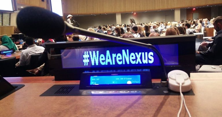 No primeiro plano a foto mostra um monitor onde se lê #wearenexus. Ao fundo auditório lotado e uma pessoa no palco, de chapéu de cowboy, fala ao microfone