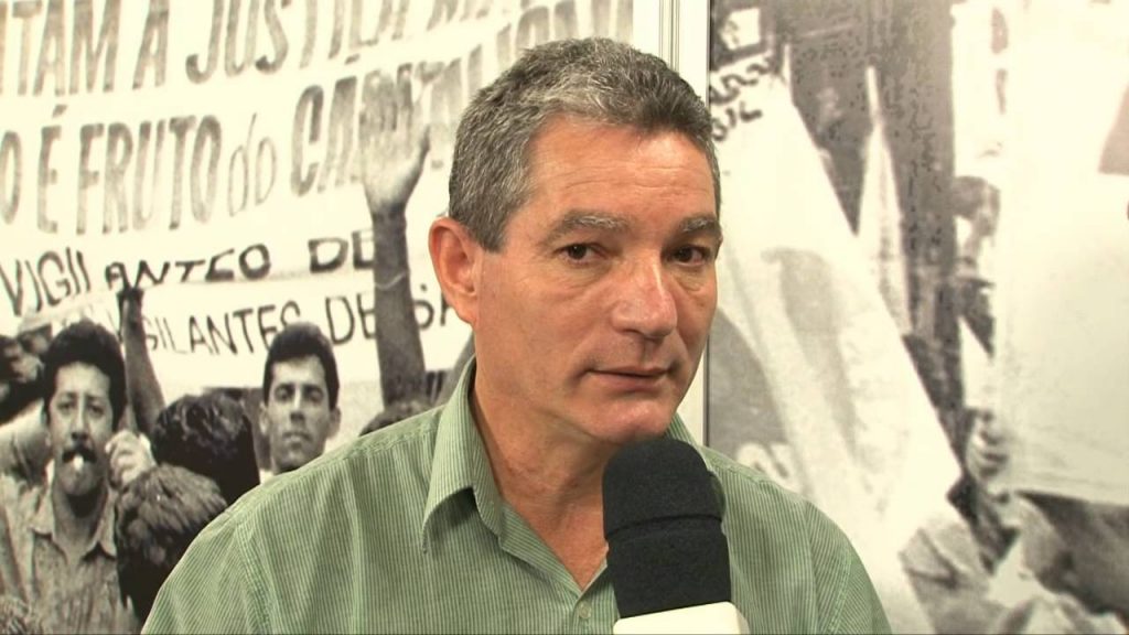 Descrição da Imagem: Carlos Clemente aparece em frente a um painel com imagens de trabalhadores. Ele tem a pele clara, cabelos grisalhos, usa camisa verde e fala ao microfone