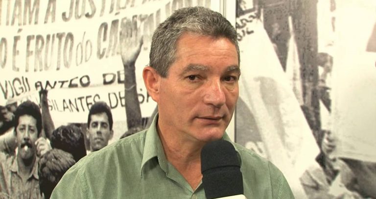 Descrição da Imagem: Carlos Clemente aparece em frente a um painel com imagens de trabalhadores. Ele tem a pele clara, cabelos grisalhos, usa camisa verde e fala ao microfone
