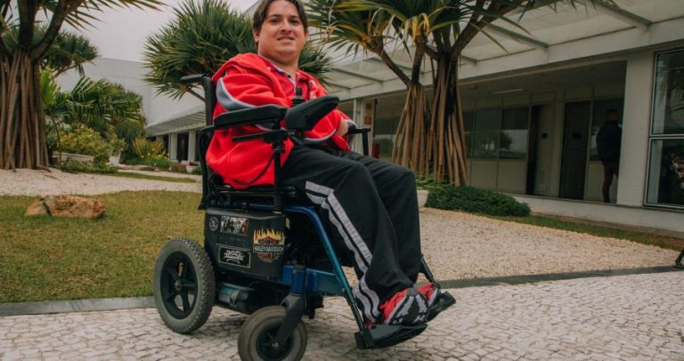 Cláudio está em uma cadeira de rodas. Ele não tem parte dos braços. É moreno, tem a pele clara e cabelos lisos e castanhos. Usa agasalho vermelho e preto